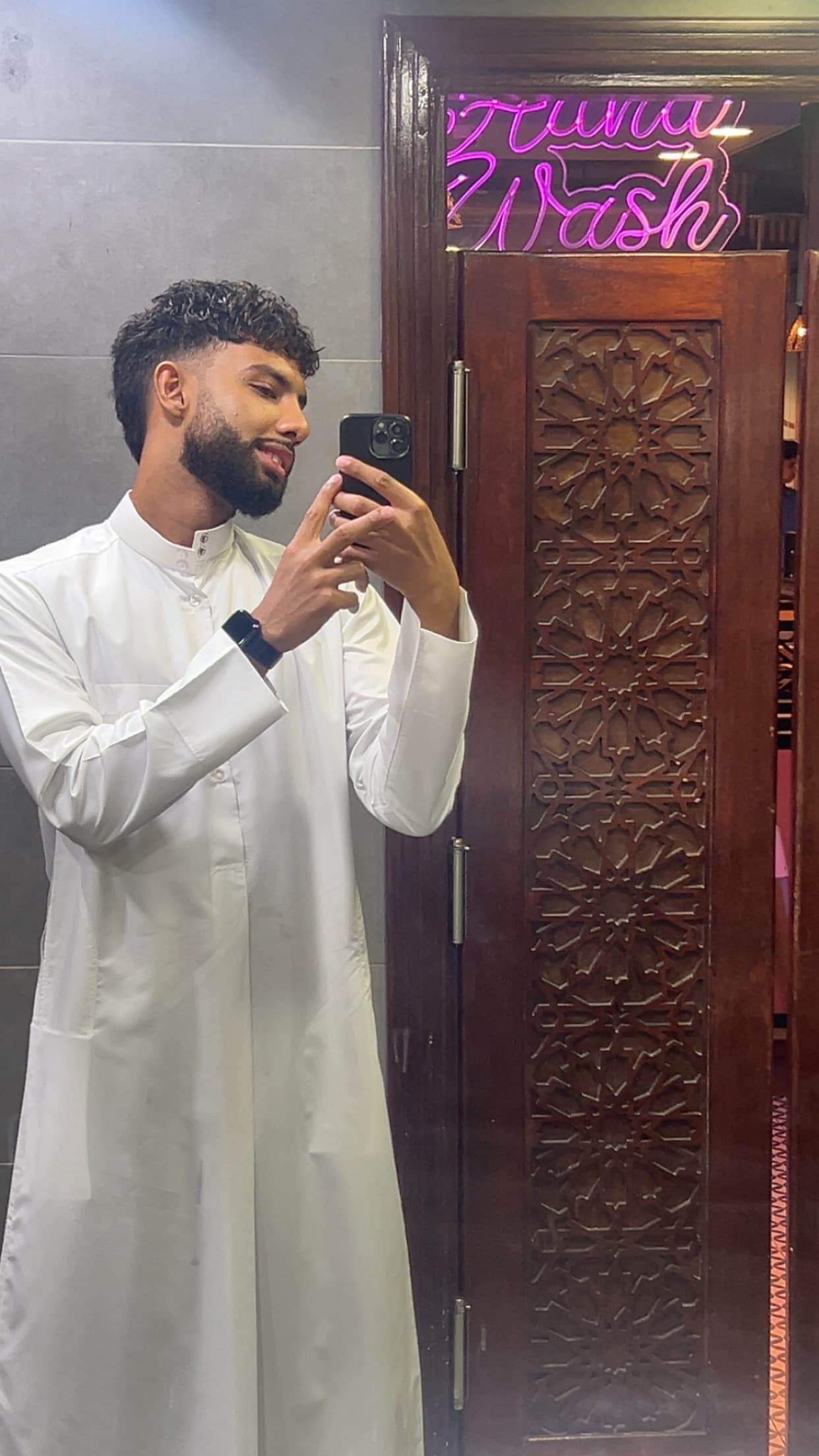 Bearded man in white thobe takes a mirror selfie beside an intricately carved wooden door.