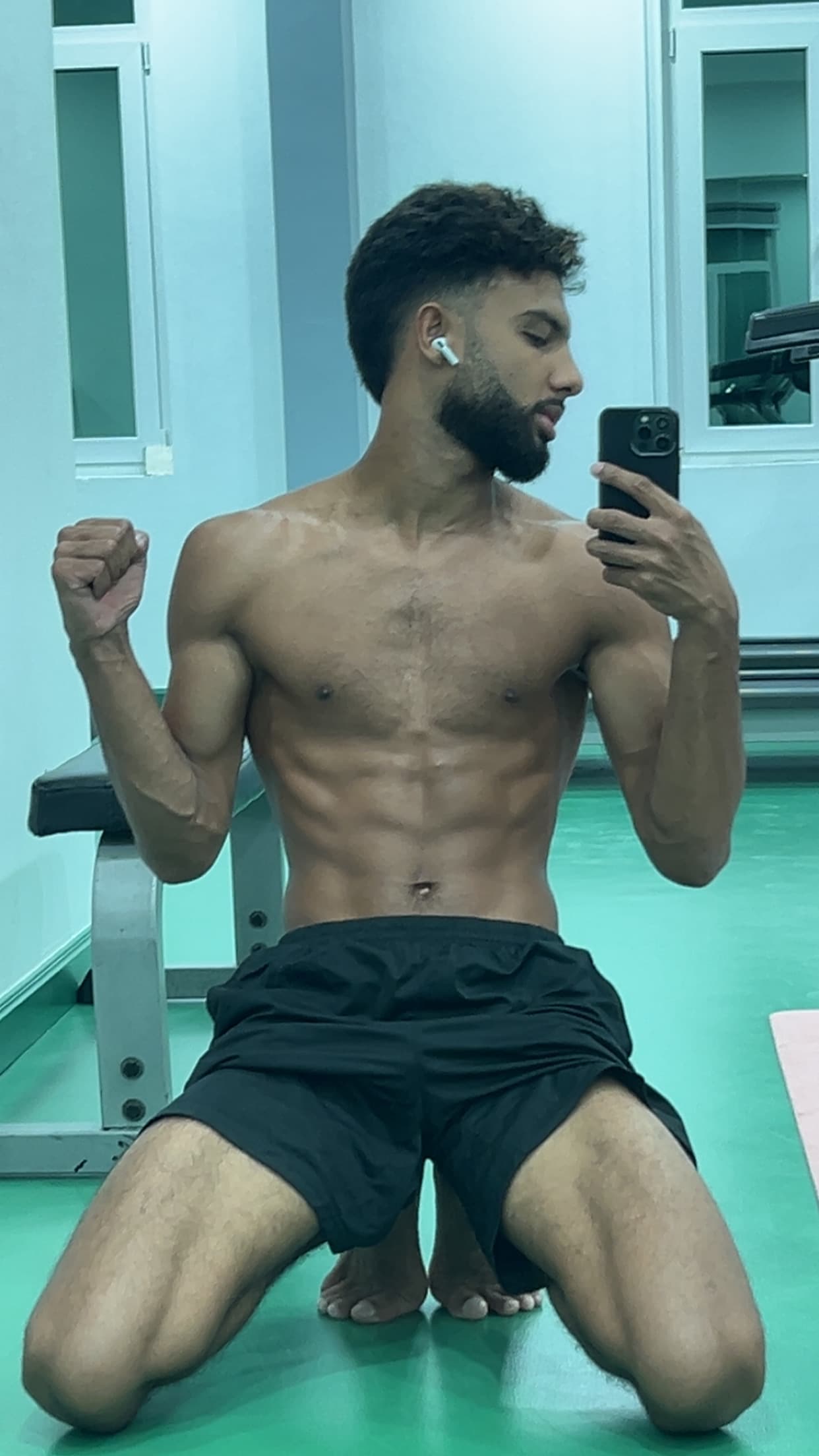 Shirtless bearded man with defined abs flexing for a mirror selfie in a gym.
