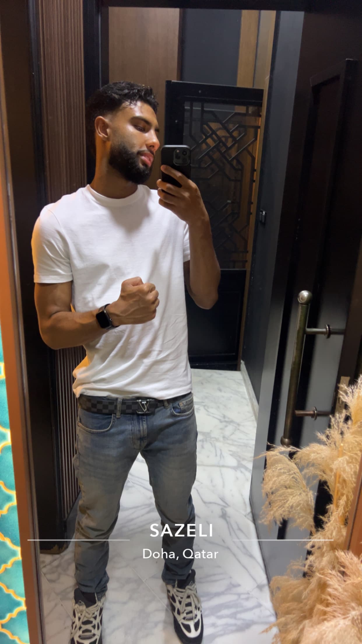 Bearded man in white t-shirt and jeans taking a mirror selfie in a modern interior.
