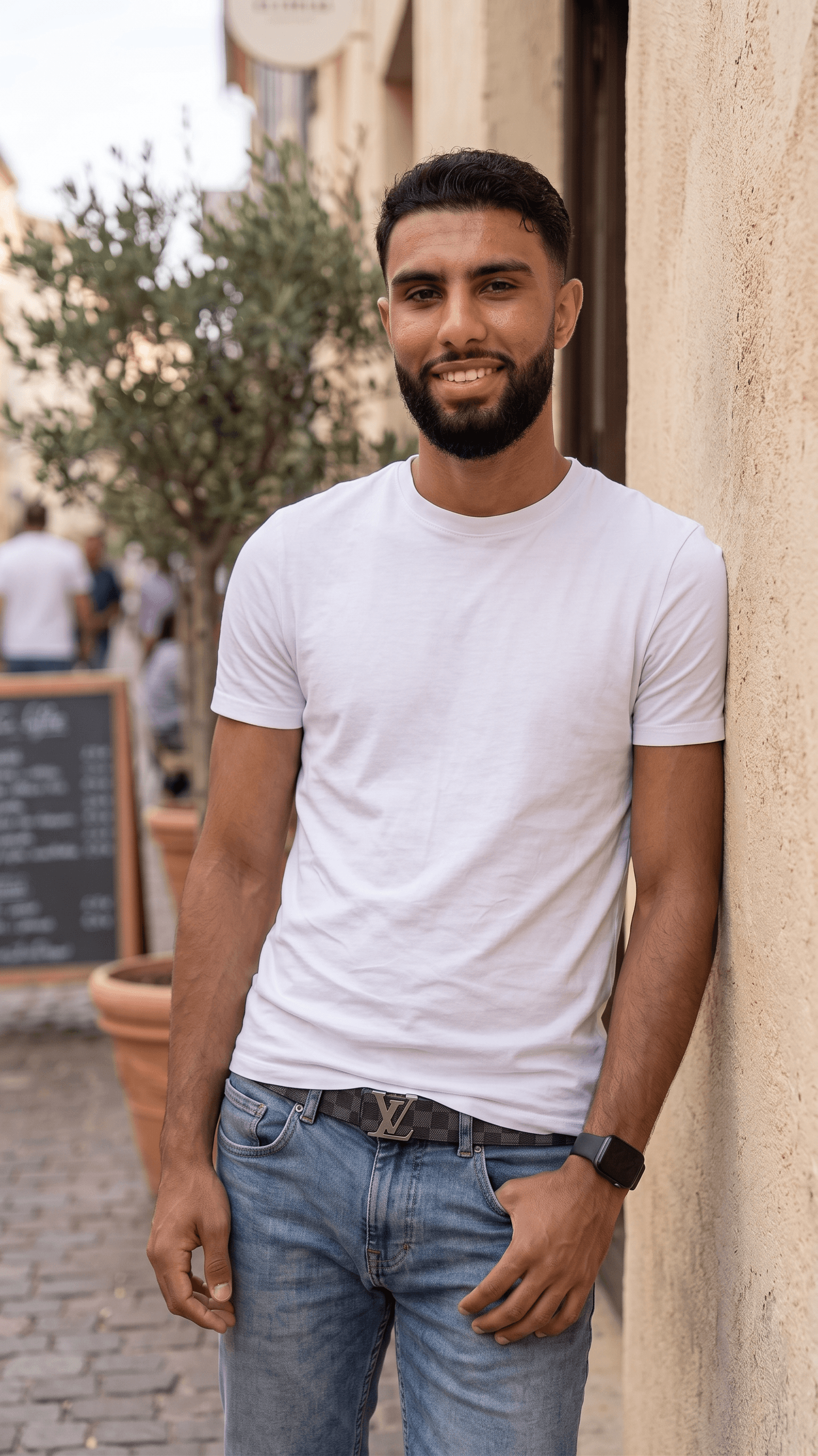 Smiling bearded man in a white t-shirt and blue jeans leaning against a stone wall.