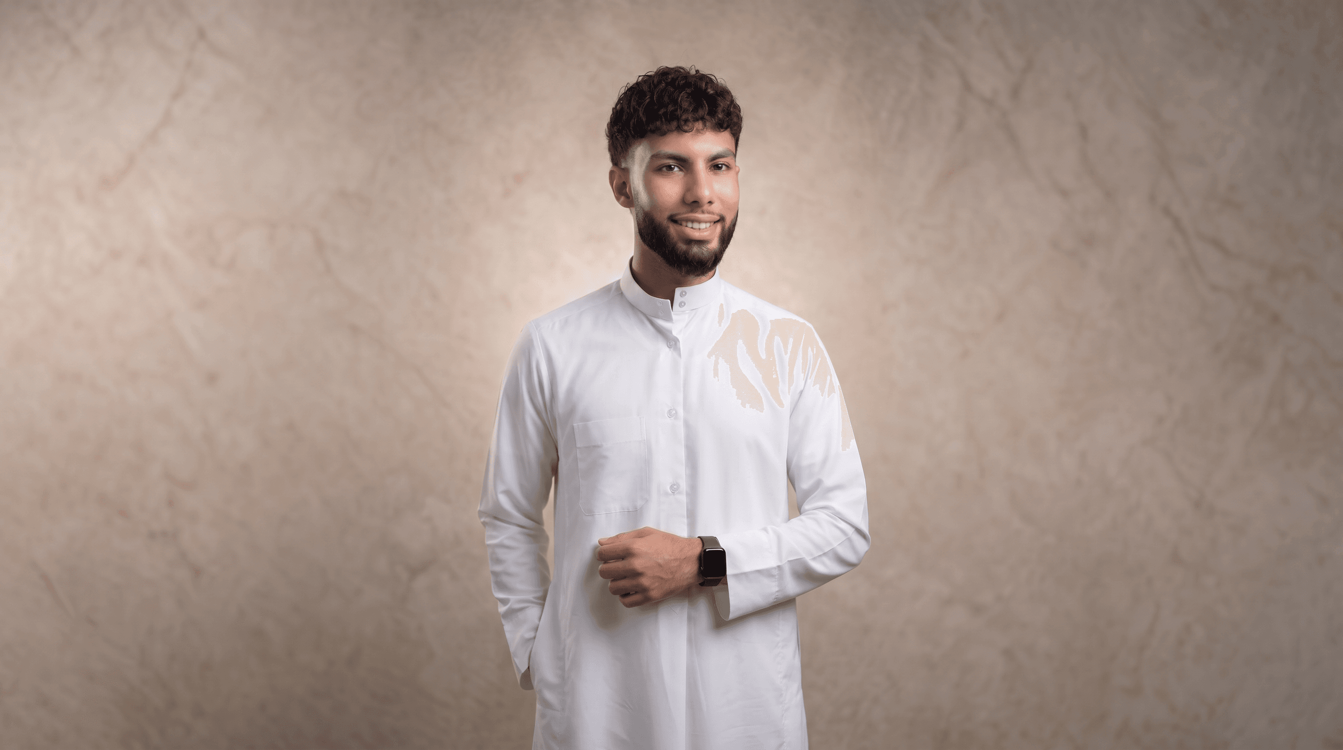 Smiling young man with a beard wearing a white thobe against a textured tan backdrop.