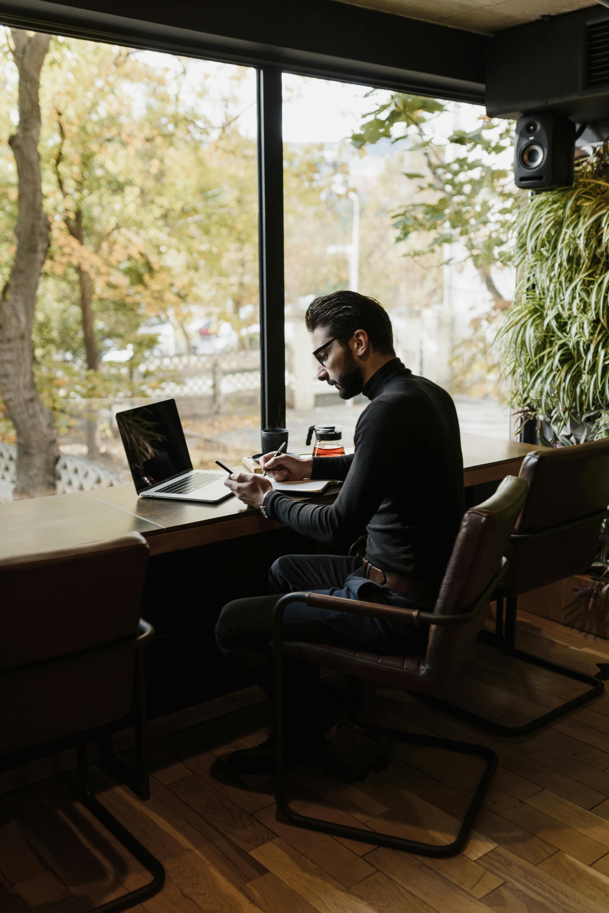Man in black turtleneck writing in a notebook at a cafe window with a laptop.