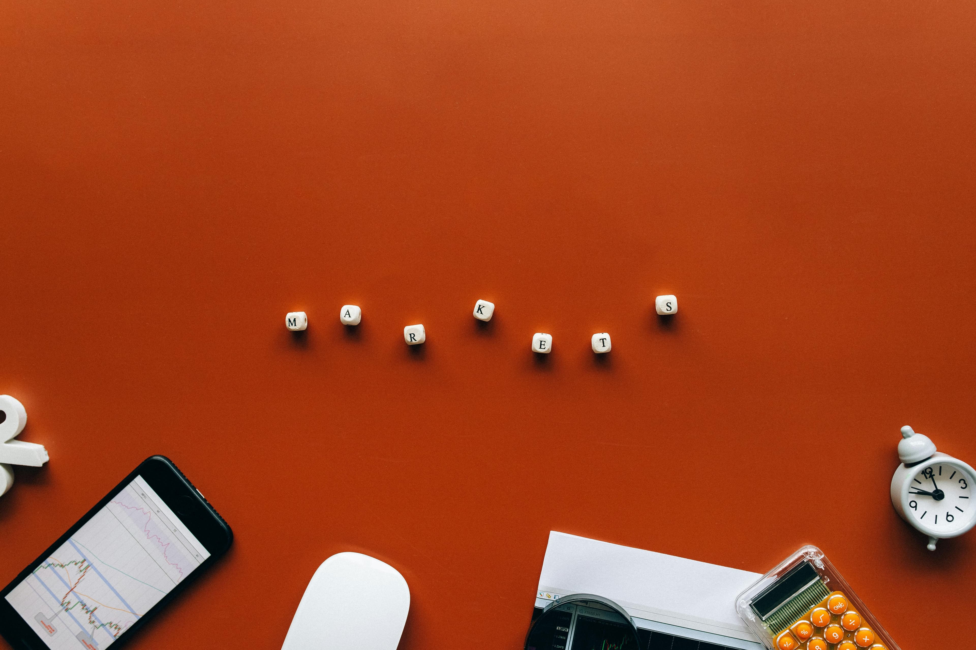Wooden blocks spelling MARKETS on orange background with a smartphone chart, calculator, and alarm clock.