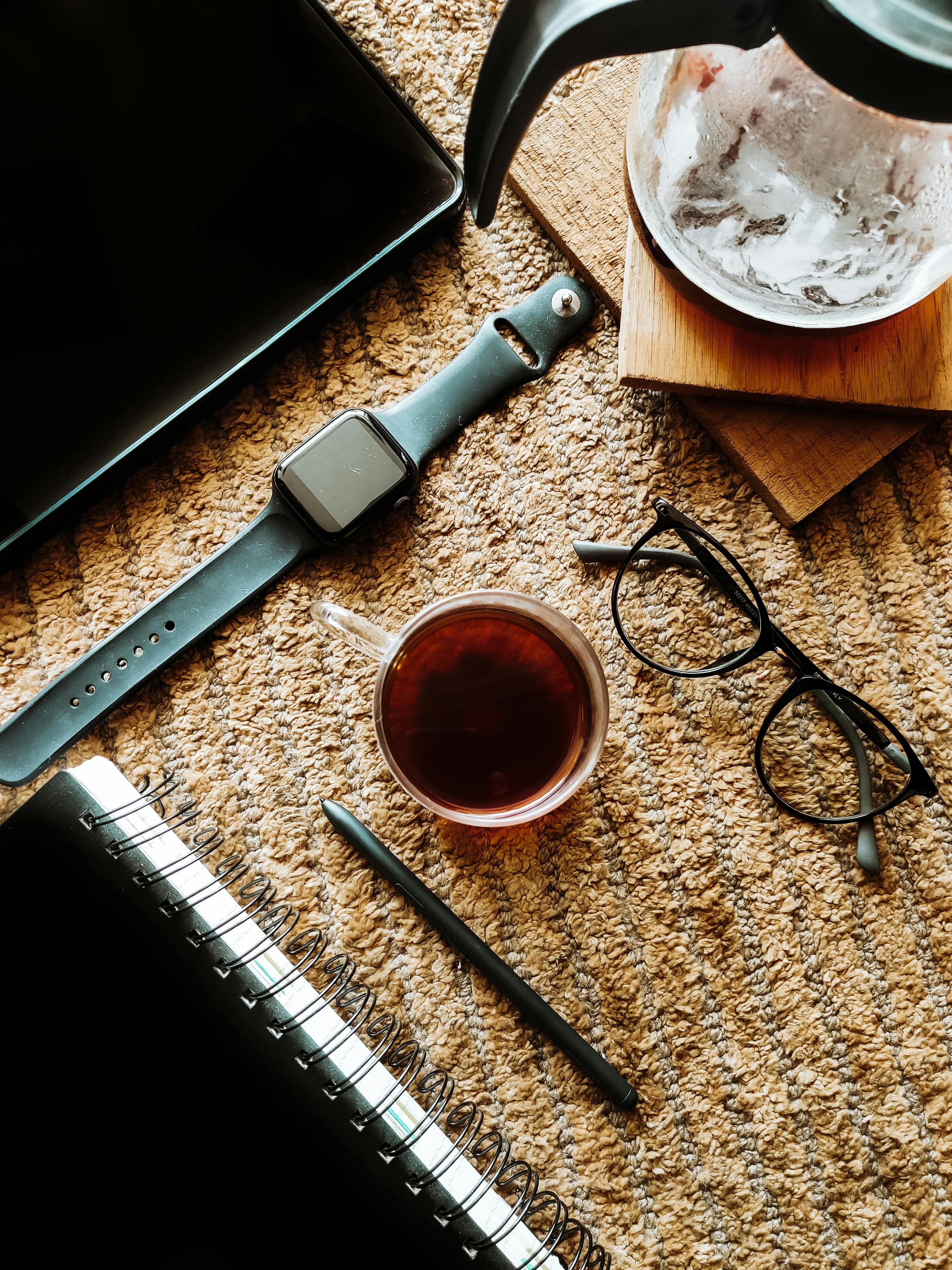 Tablet, smartwatch, cup of tea, glasses, and notebook on a beige textured carpet.
