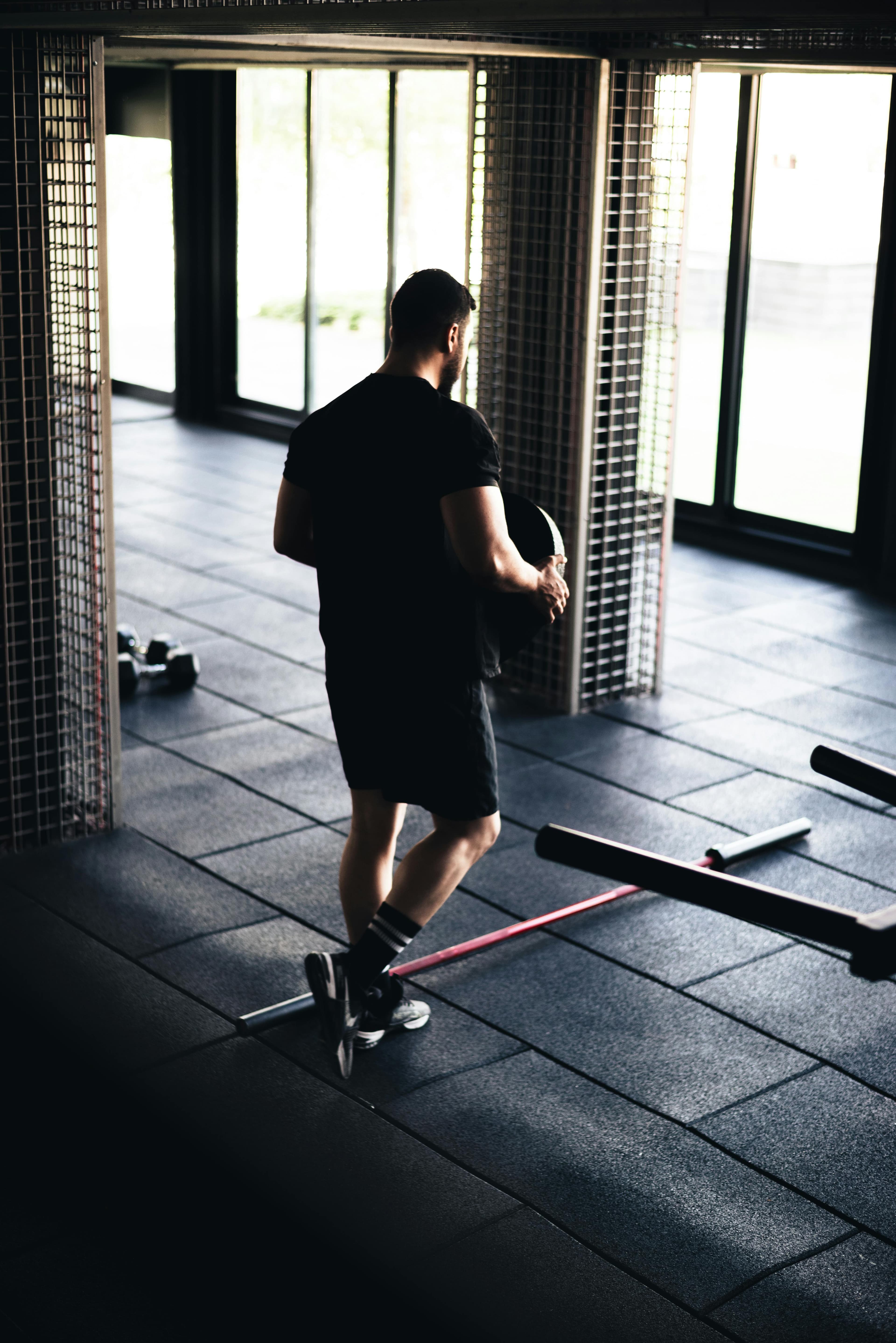 Man in black sportswear carries a weight plate across a sunlit, modern gym floor.