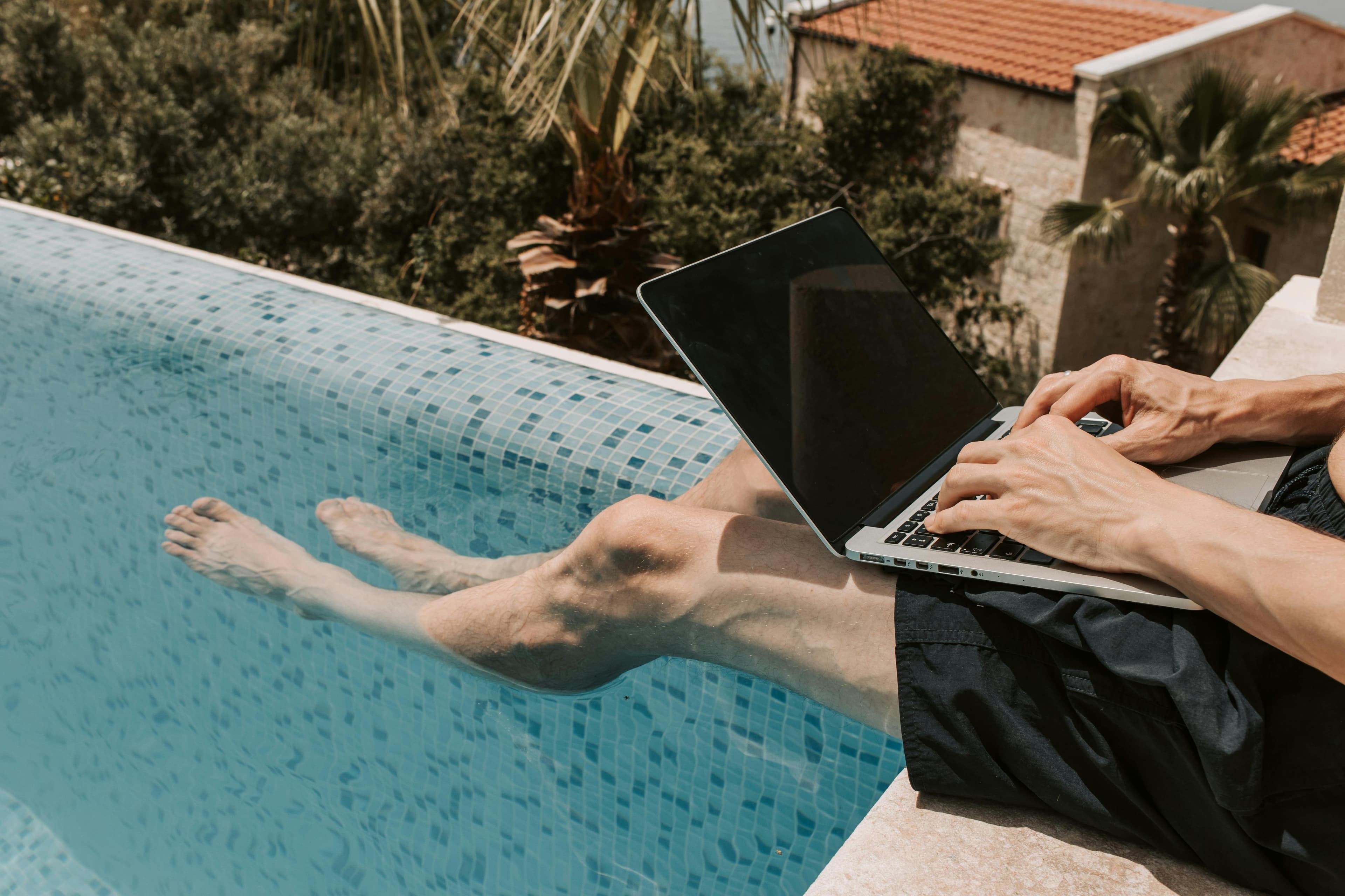 Person working on a laptop while dangling their legs in a bright blue swimming pool.